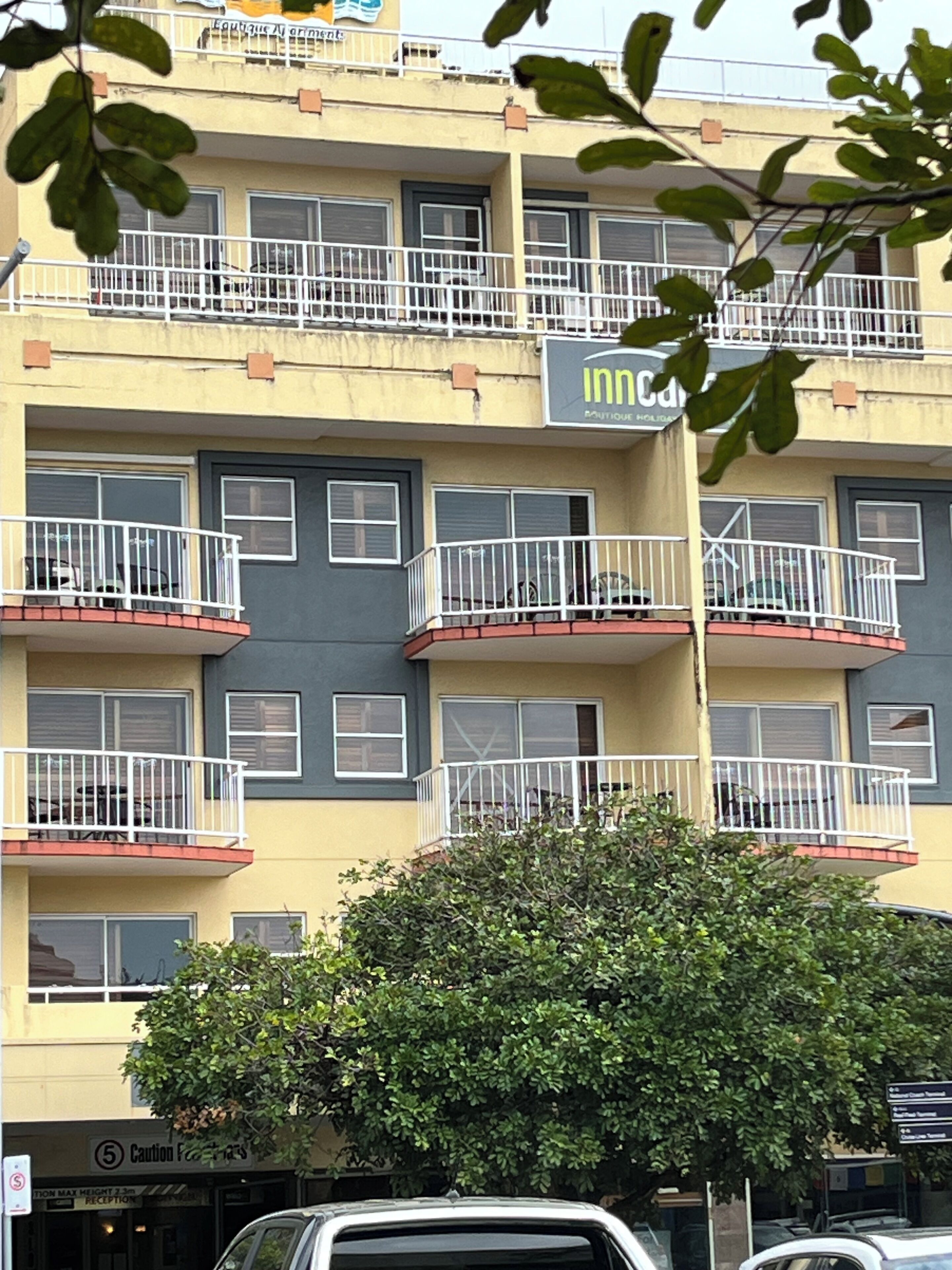 A multi-story building with balconies, a visible storefront with a sign, and a tree in the foreground.