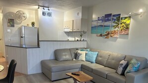 Una sala moderna con un sofá de cuero gris, una mesa de centro de madera y una silla negra. Se ve un área de cocina con armarios blancos y un refrigerador al fondo.