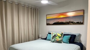 Un dormitorio con cama, ventilador de techo, un cuadro de un atardecer y cortinas.
