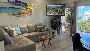 Una sala de estar con un sofá de cuero color beige, una mesa de centro y un televisor montado en la pared. Se observan tres cuadros enmarcados, una planta en maceta y un balcón a través de la ventana.