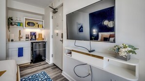 A compact hotel room with a kitchenette, a flat-screen TV mounted on the wall, and a small shelf with a decorative plant. - Ocean View Efficiency, Newly Renovated,Jacuzzi Tub (Myrtle Beach)