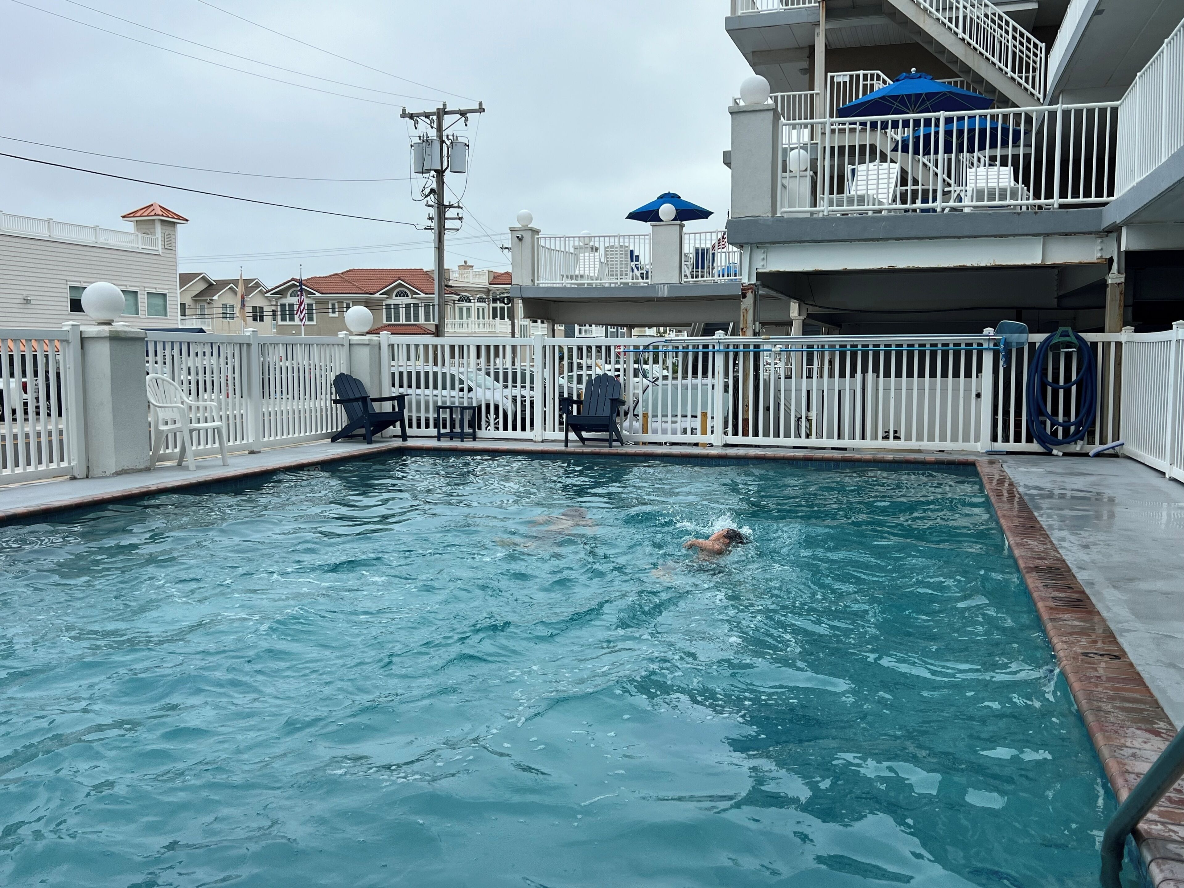 Una piscina con una persona nadando, rodeada por una valla blanca, y un edificio con un balcón al fondo.