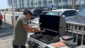 A man grilling at an outdoor barbecue station with a Genesis grill, surrounded by parked cars and a multi-story building in the background. - Beachfront Condo (Summer Sands #214) with outdoor pool & BBQ grills! (North Wildwood)
