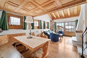Een ruime kamer met een houten plafond, een eettafel met stoelen, een bank en een blauwe fauteuil.