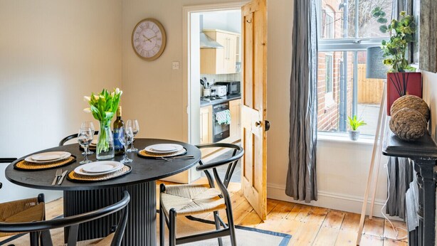 A dining area with a round table set for four, chairs, a clock on the wall, and a view of the kitchen through an open door. - Golden Triangle 2 bed + Garden + Parking (Norwich)