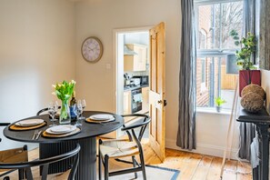 Un coin repas avec une table ronde dressée pour quatre personnes, des chaises, une horloge au mur et une vue sur la cuisine par une porte ouverte.