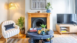 A living room with a striped armchair, a round coffee table with books and cups, a fireplace, a TV, and a large window. - Golden Triangle 2 bed + Garden + Parking (Norwich)