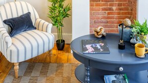 A living room with a striped armchair, a round coffee table, a floor lamp, a bookshelf, and a brick fireplace. - Swift Host |Off Unthank Road | 2 bed + Parking + Garden (Norwich)