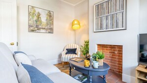 A living room with a sofa, armchair, coffee table, bookshelf, fireplace, and a painting on the wall. - Swift Host |Off Unthank Road | 2 bed + Parking + Garden (Norwich)