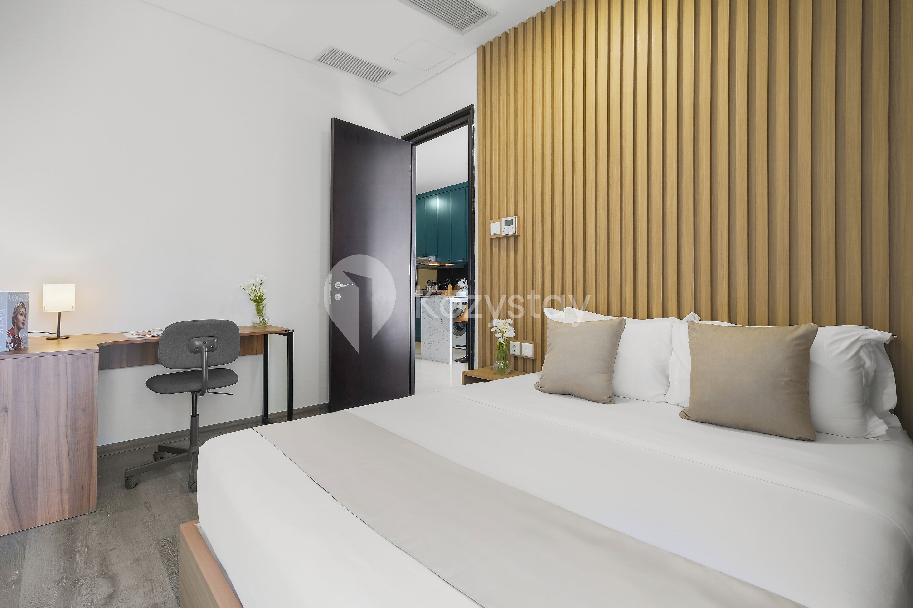 A modern hotel room with a large bed, a desk with a chair, a wooden accent wall, and a view into the kitchen area.