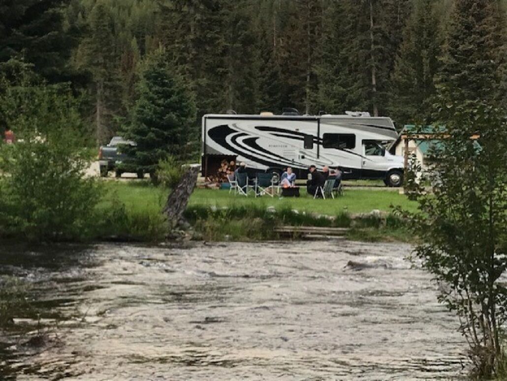 A white RV parked by a river with a forest in the background.