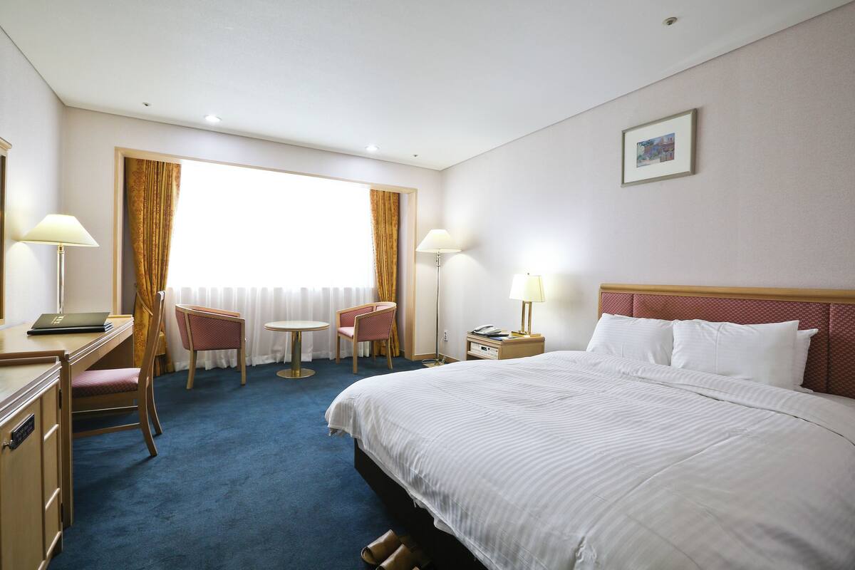 A hotel room with a large bed, a desk, a chair, a small table, and a window with curtains.