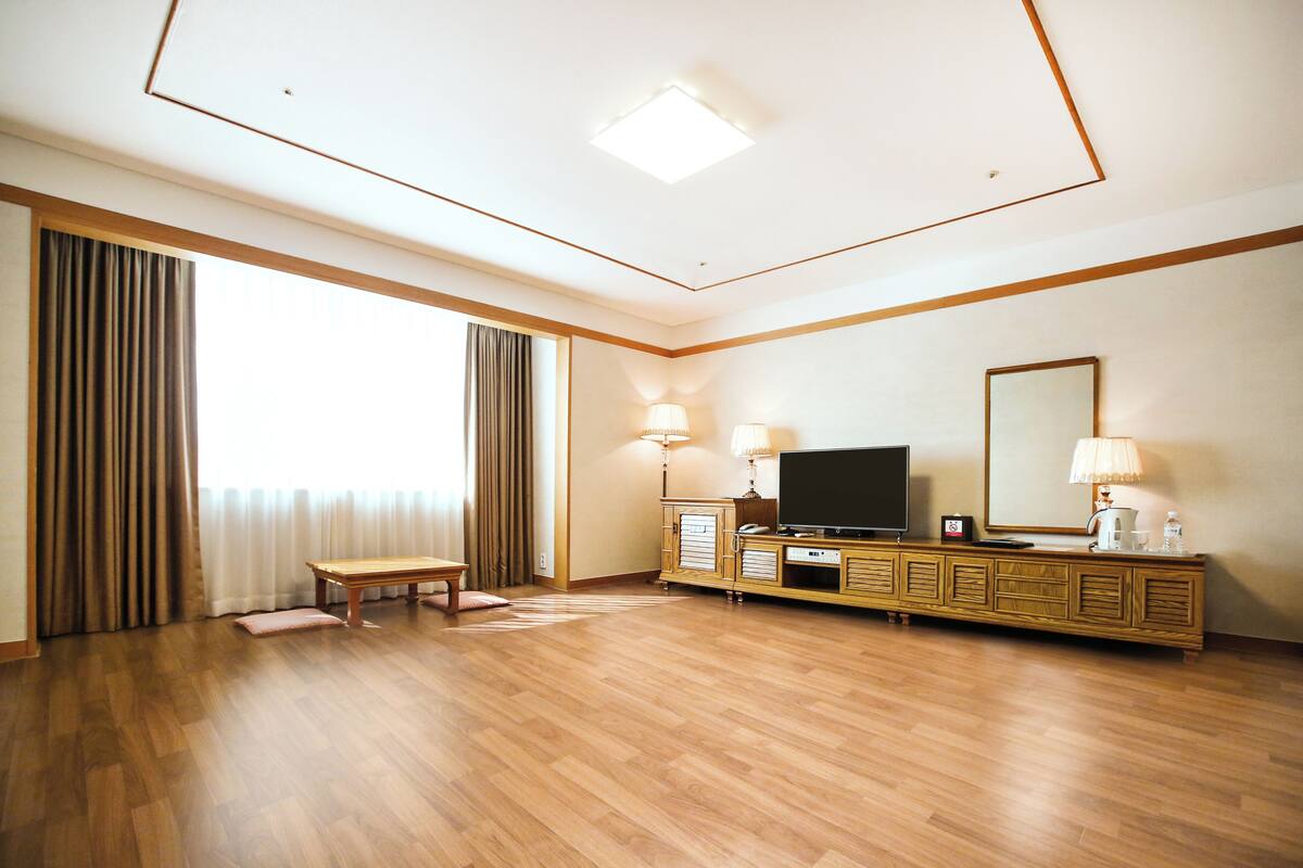 A spacious hotel room with a large window, a flat-screen TV, a wooden cabinet, and a bench.