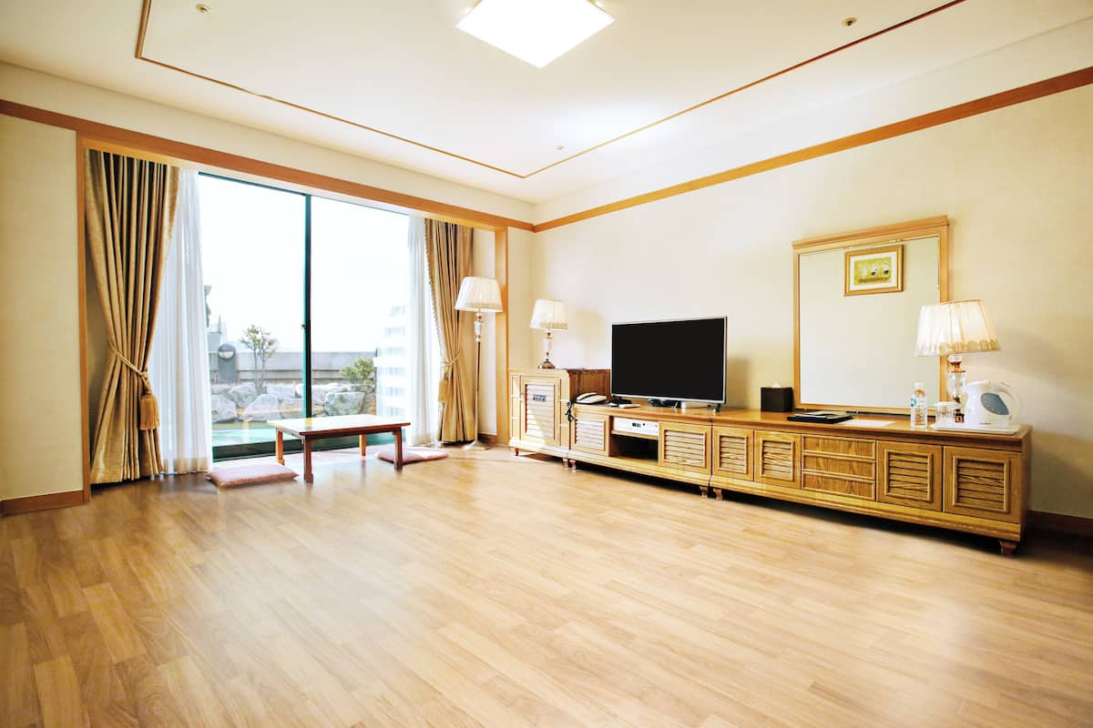 A spacious hotel room with a large sliding glass door leading to a balcony, a wooden TV stand with a flat-screen TV, and a large mirror.