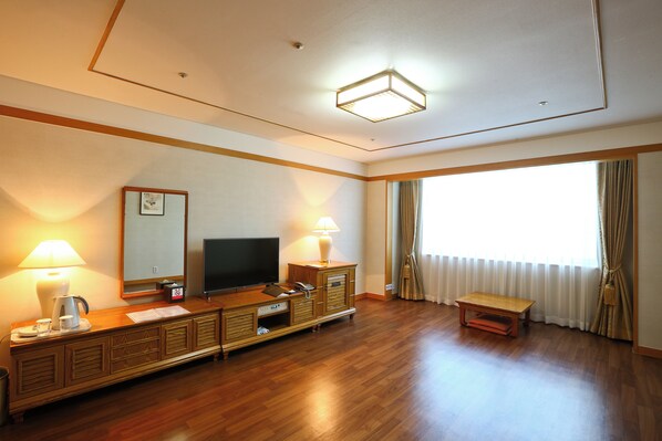 A hotel room with a wooden floor, a TV on a wooden cabinet, a lamp, a small table, and a window with curtains. - Legend Hotel (Daejeon)