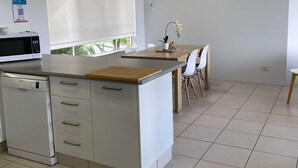 A modern kitchen with white cabinetry, a built-in microwave, and a dishwasher. There is a dining area with a wooden table and chairs. - U07 Tripcony 2 Bedroom w Balcony (Sunshine Coast)