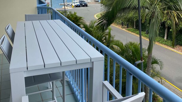 A balcony with a white table and chairs, overlooking a coastal road with palm trees and a view of the sea. - U07 Tripcony 2 Bedroom w Balcony (Sunshine Coast)