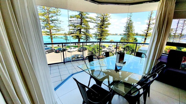Un balcon avec une table et des chaises en verre, donnant sur une plage et des arbres.