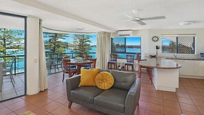 A modern living room with a grey sofa, yellow cushions, a dining area with wooden chairs, a kitchen with a white countertop, and a balcony with a view of the sea.