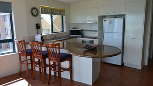 A compact kitchen with a central island, white cabinets, a microwave, and a refrigerator. There are three wooden chairs with blue cushions around the island.