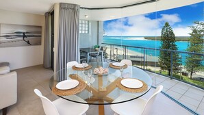Un comedor moderno con mesa de vidrio y sillas blancas, con vistas a una playa y al océano.