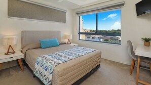Un dormitorio con cama, escritorio, silla, ventana con vistas, ventilador de techo y televisor en la pared.