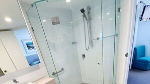 A modern bathroom with a glass-enclosed shower, white vanity, and a blue chair in the background.