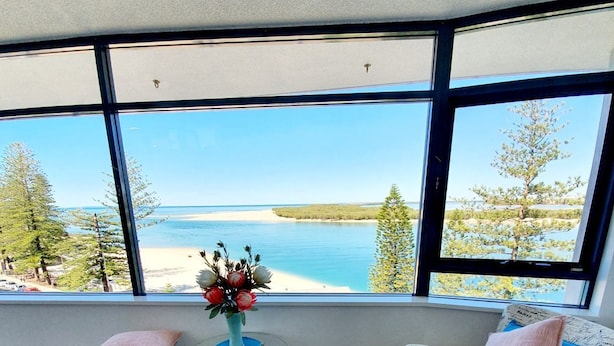 A room with a large window overlooking a beach and ocean. There is a small round table with a vase of flowers, a patterned armchair, and a cushion.