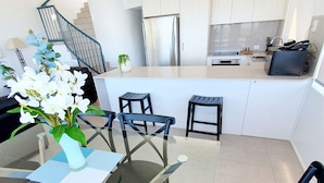 A modern kitchen with white cabinets, a stainless steel refrigerator, and a small dining area with a glass table and black chairs.