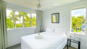 A hotel room with a large bed, a ceiling fan, a nightstand, a lamp, and a window with a view of palm trees.