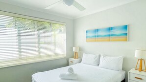 Une chambre d’hôtel avec un lit, un ventilateur de plafond, une fenêtre avec des stores, une table de chevet avec une lampe et un tableau au mur.