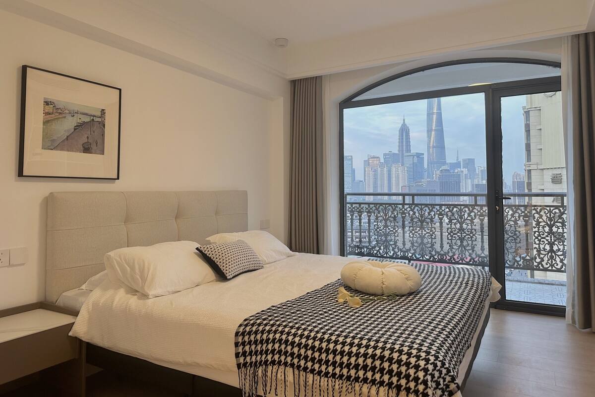 A bedroom with a bed, a cityscape view through a large window, and a framed picture on the wall.