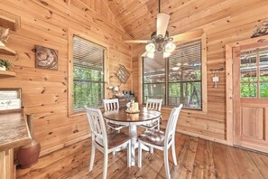 A wooden cabin interior with a round dining table, white chairs, a ceiling fan, and large windows.