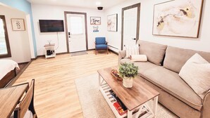 A modern living room with a sofa, a coffee table with a plant, a TV mounted on the wall, and a door with a window. - Cozy Studio Cottage with Harbor View/Spacious Full Kitchen & Pet Friendly (Bodega Bay)