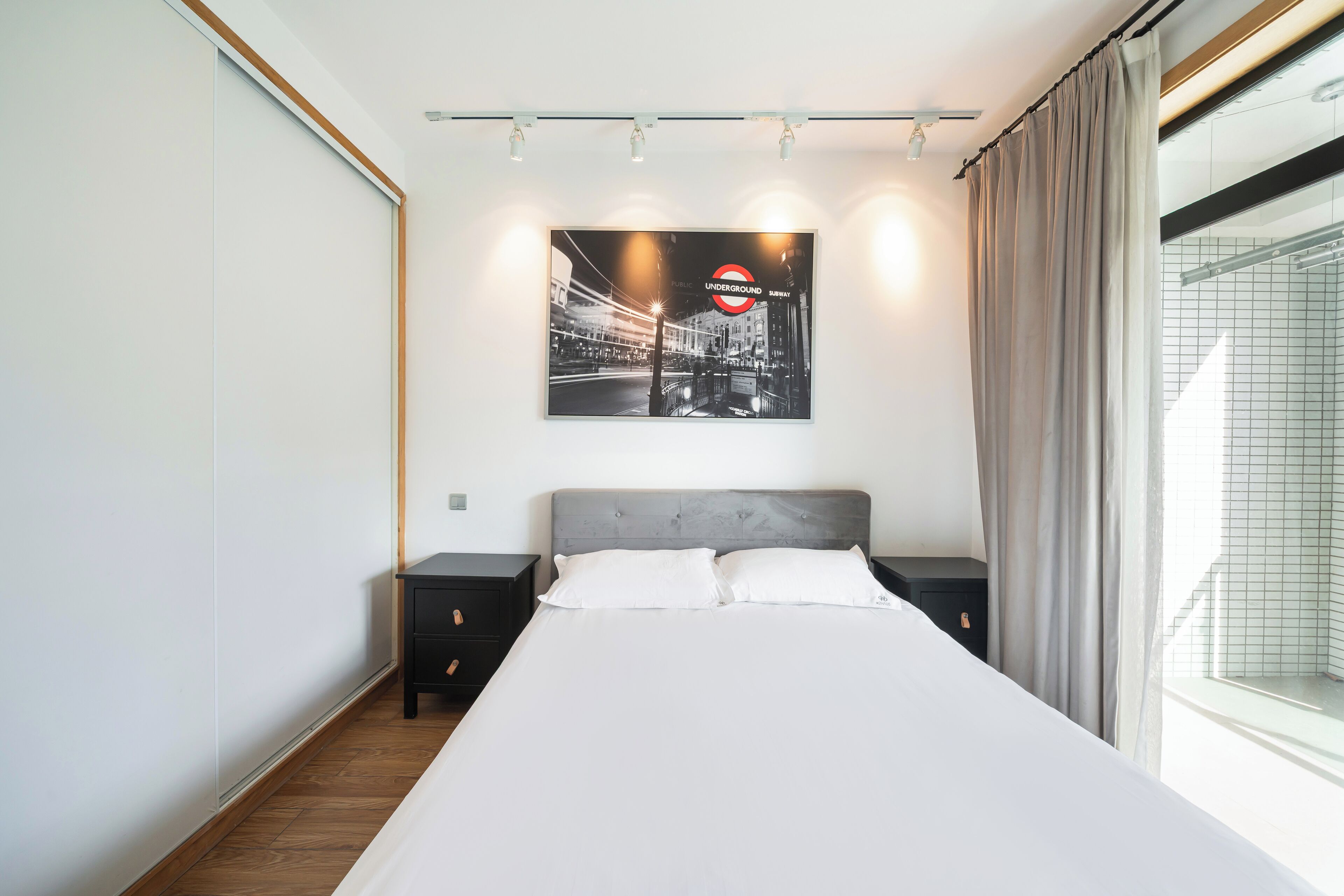 Un dormitorio moderno con una cama grande, una mesita de noche, una lámpara de pared y un ventanal con cortinas.
