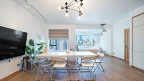 A modern living room with a large flat-screen TV, a dining table with chairs, a city view through large windows, and a stylish pendant light.