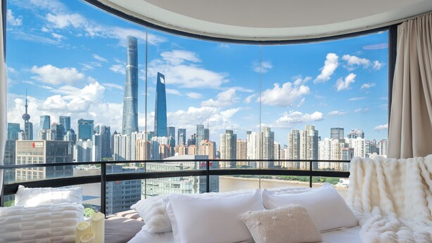 A hotel room with a large window offering a city skyline view, a neatly made bed with white linens, and a small table with towels and a bottle.