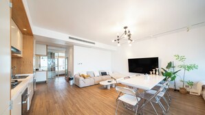 A modern kitchen with wooden cabinets, a dining area with a white table and chairs, a sofa, and a TV mounted on the wall.