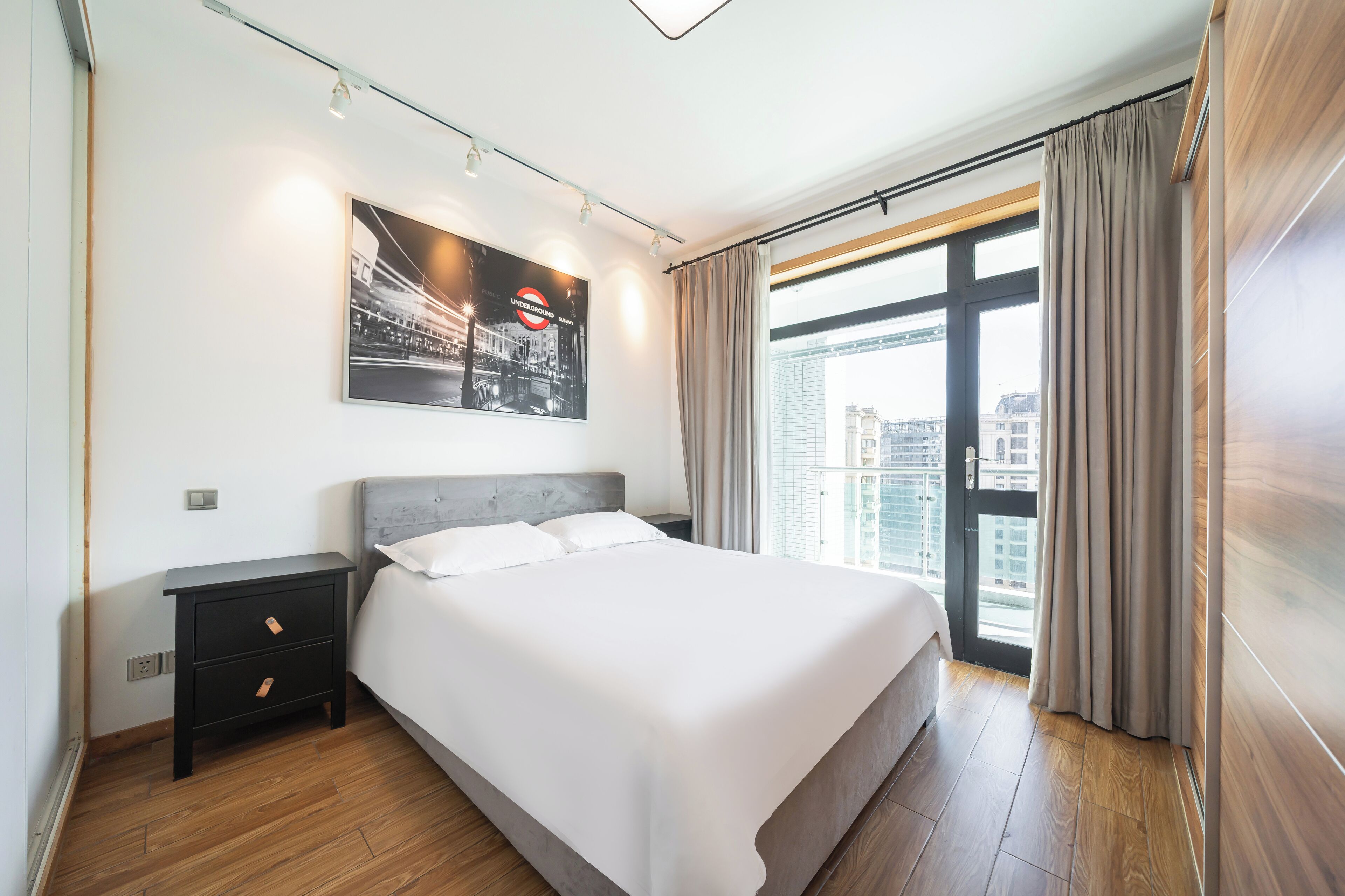 A modern bedroom with a large bed, a nightstand, a wall-mounted light, a cityscape view, and a window with curtains.