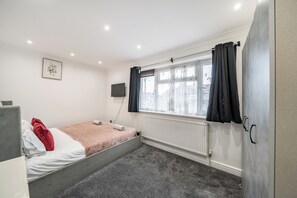 A single-bed room with a grey carpet, a white radiator, a mounted TV, and a window with black curtains.