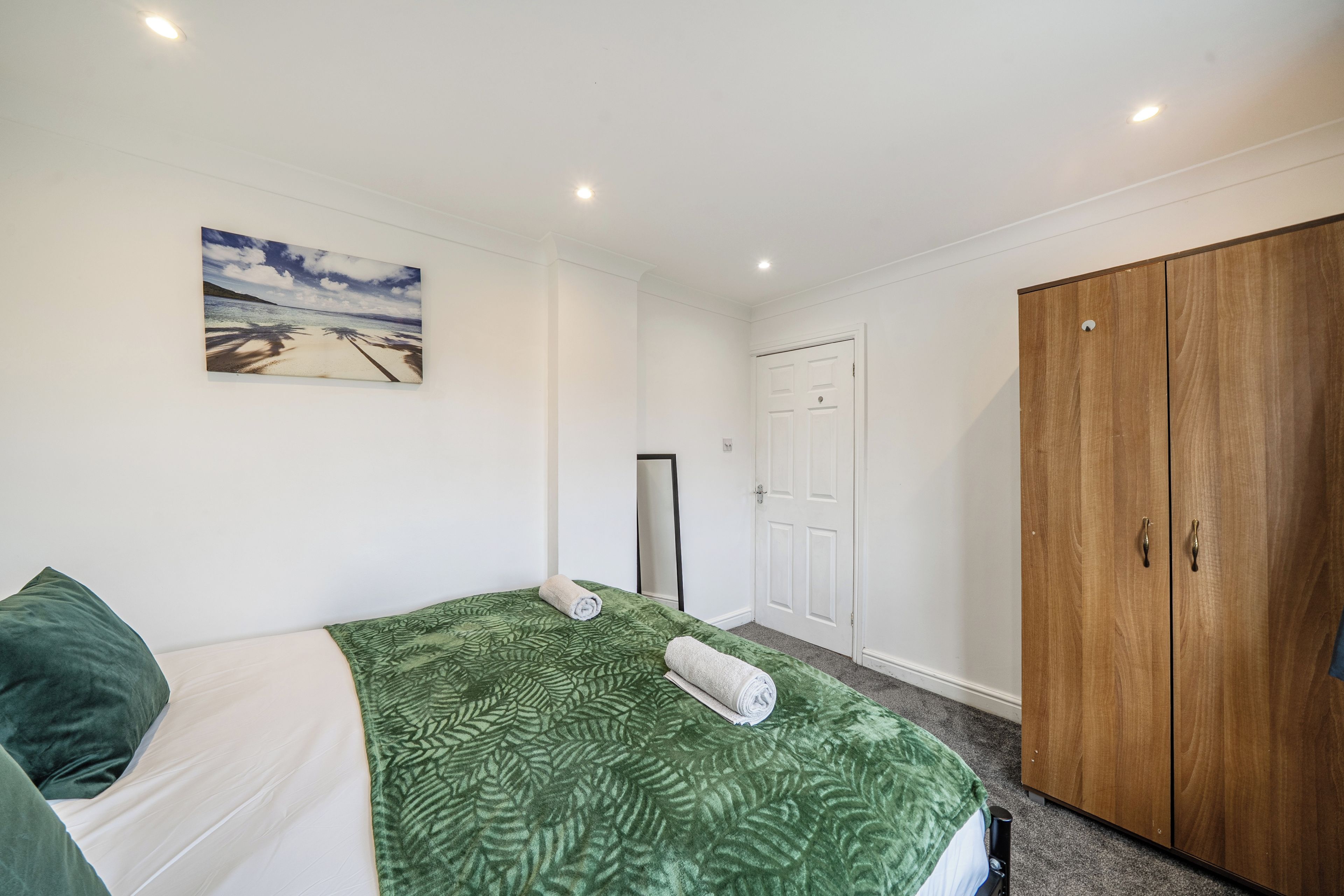 A bedroom with a bed, a green patterned bedspread, a painting of a beach scene, a wardrobe, and a door.