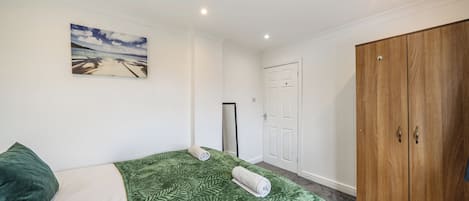A bedroom with a bed, a green patterned bedspread, a painting of a beach scene, a wardrobe, and a door.