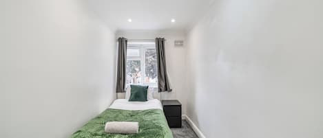 A narrow, well-lit room with a single bed, a radiator, and a window with curtains.