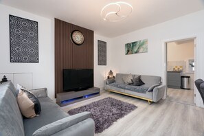 A modern living room with a grey sofa, a flat-screen TV, decorative wall panels, and a contemporary light fixture. - Cosy Cottage - Sleeps 6, Parking & Pet Friendly! (Nelson)
