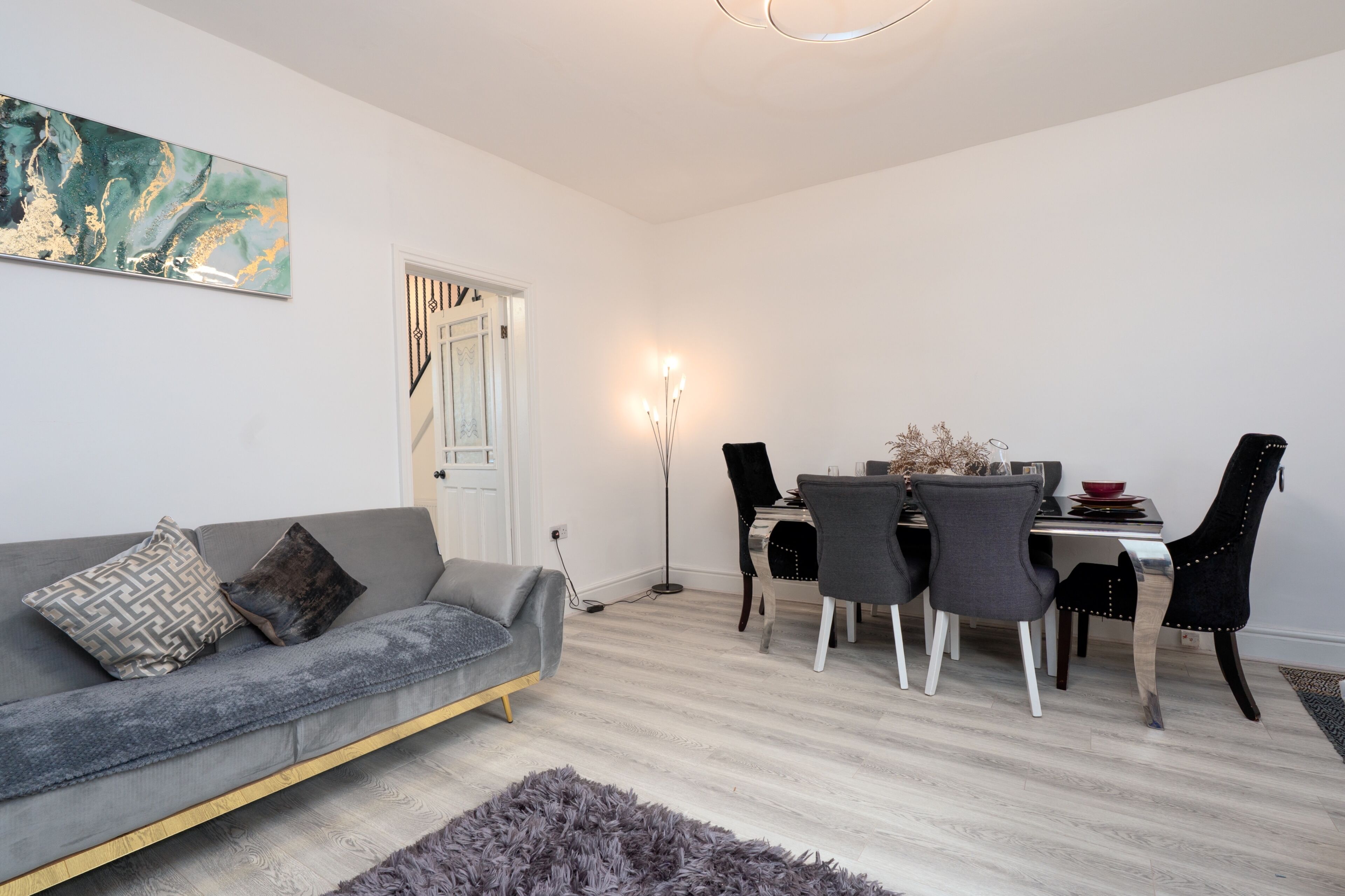 Uma sala de estar moderna com um sofá cinza, uma mesa de jantar com cadeiras, um abajur e um quadro na parede.