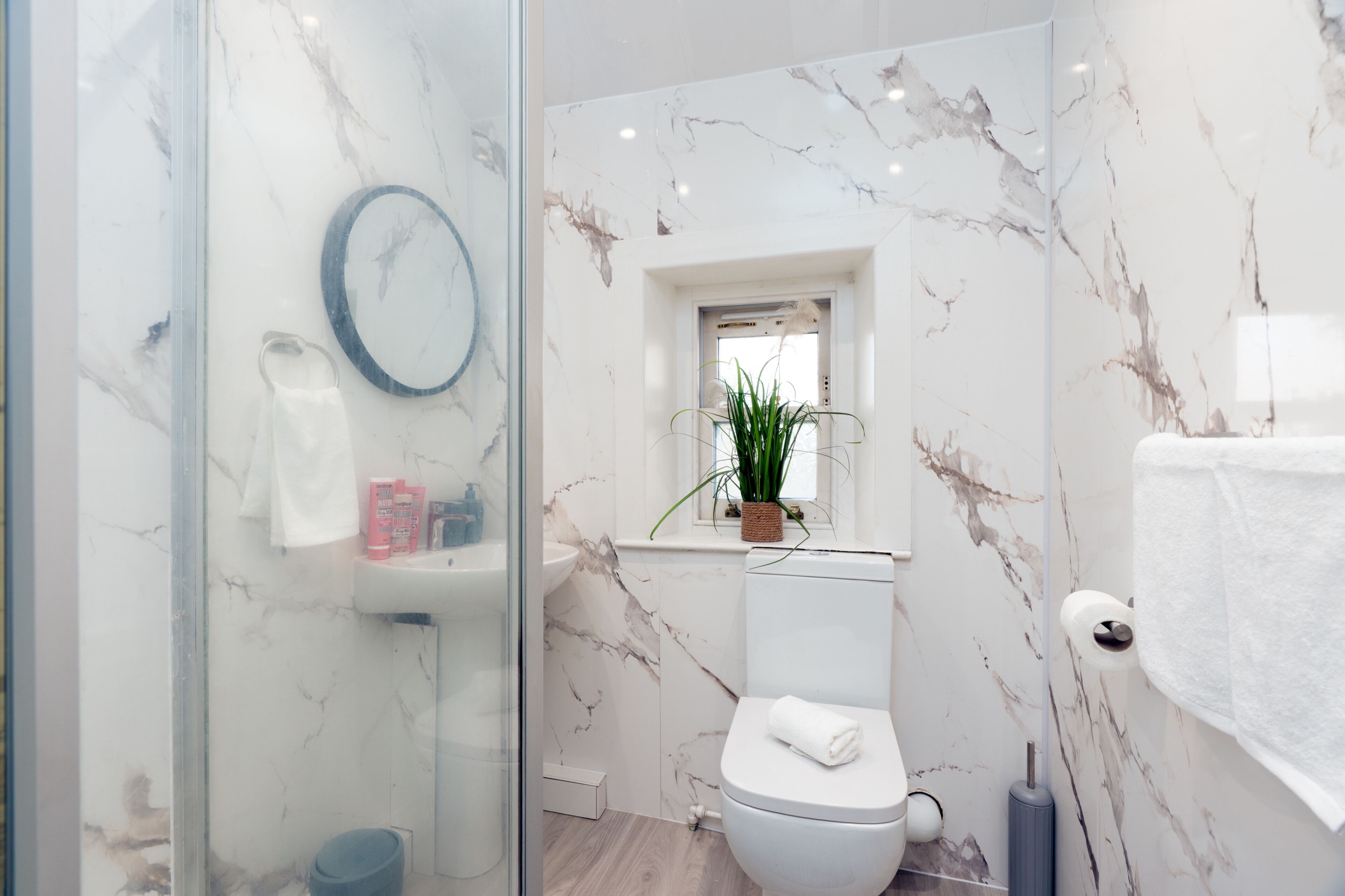 Une salle de bain moderne avec une douche à l’italienne en verre, des toilettes blanches, un pare-baignoire en marbre et une petite fen être agrémentée d’une plante.