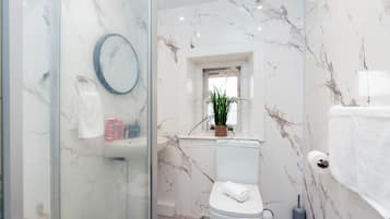 Une salle de bain moderne avec une douche à l’italienne en verre, des toilettes blanches, un pare-baignoire en marbre et une petite fenêtre agrémentée d’une plante.