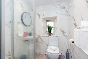 A modern bathroom with a glass shower enclosure, a white toilet, a marble backsplash, and a small window with a plant. - Cosy Cottage - Sleeps 6, Parking & Pet Friendly! (Nelson)