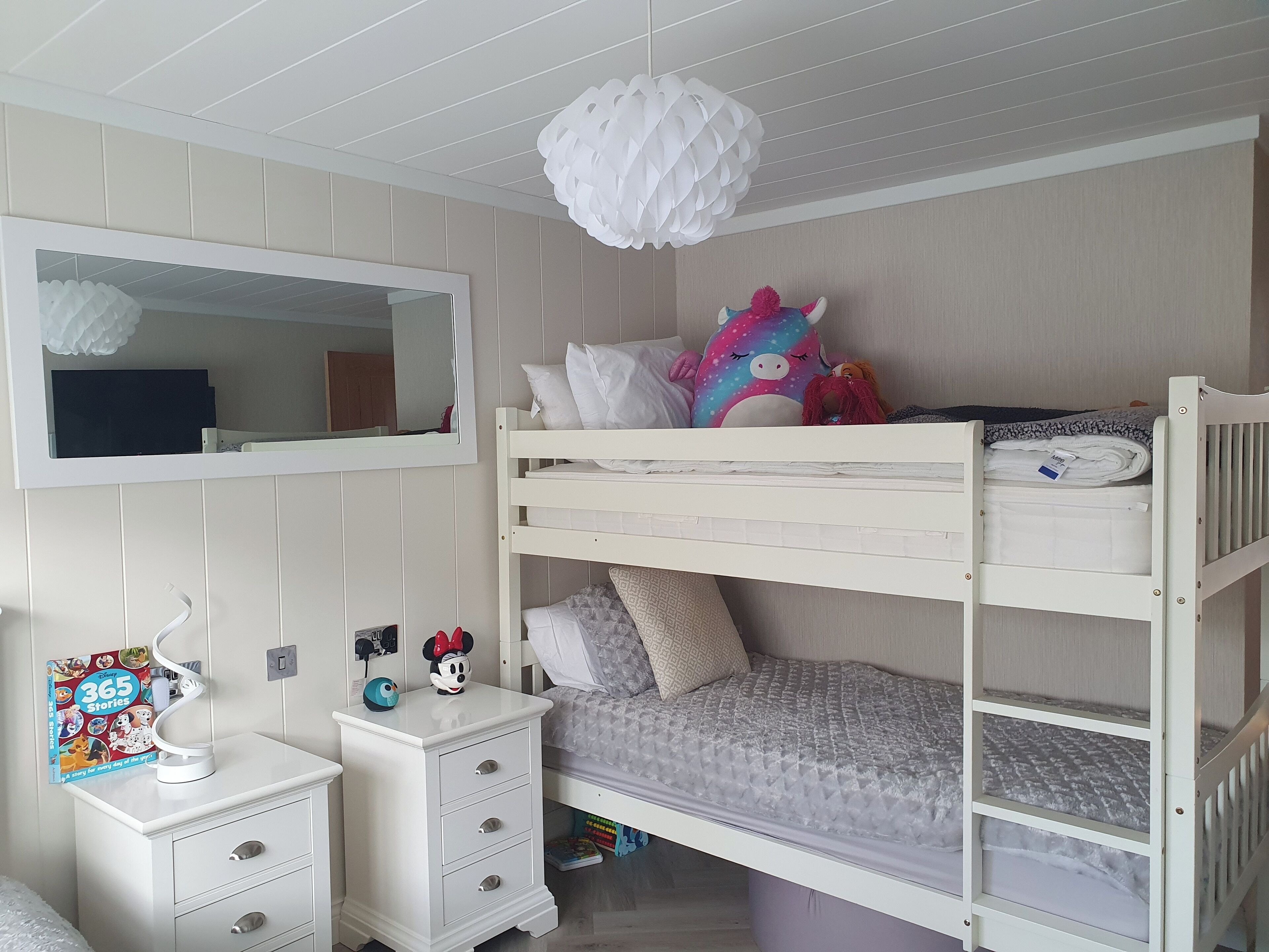A bunk bed with a white headboard, a bedside table with drawers, a mirror, and a hanging light fixture.