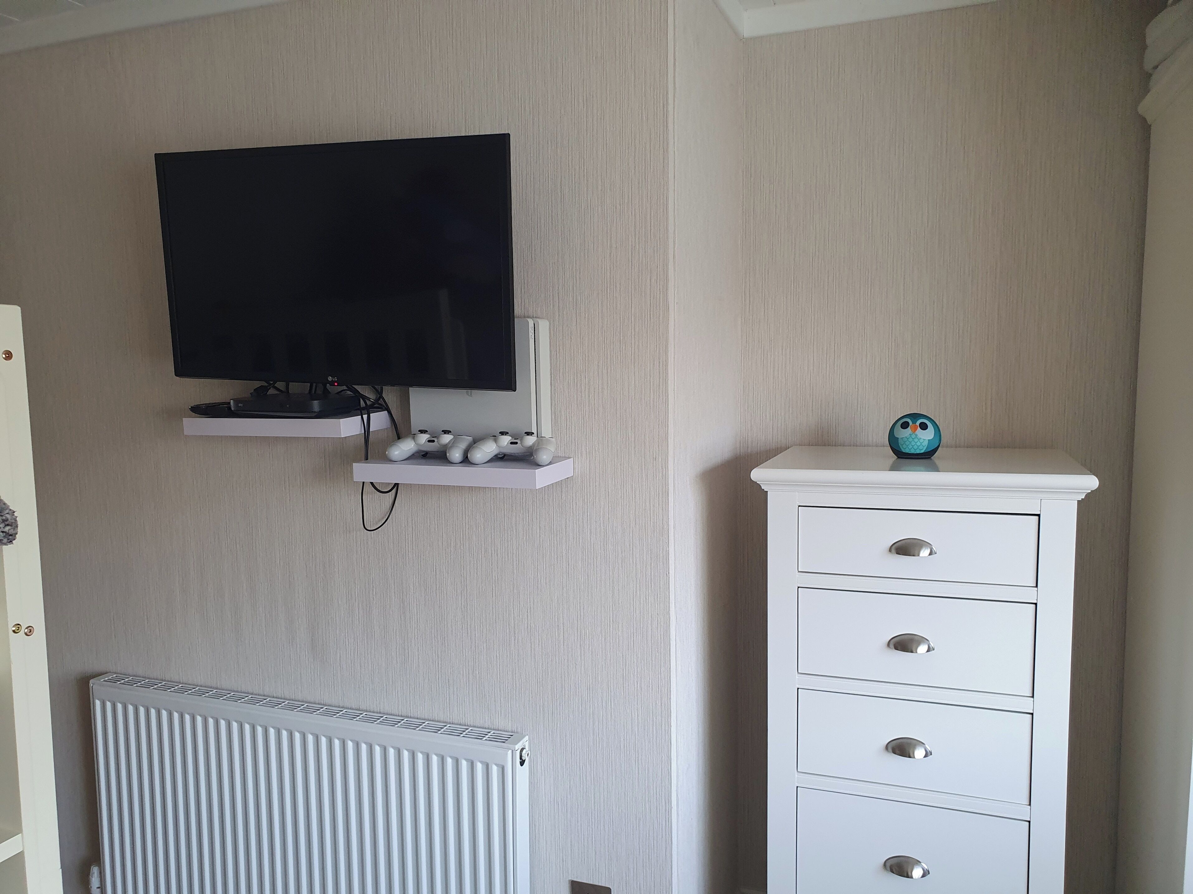 Une pièce avec un téléviseur fixé au mur, une commode blanche avec des tiroirs, un radiateur et une étagère sur laquelle est posée une chouette en jouet.